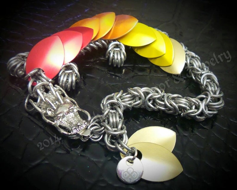 May include: A silver dragon scale chainmail bracelet with a dragon head charm. The scales are red, orange, and yellow.
