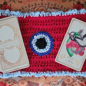 May include: Two tarot cards, one with the text "Enchanted Blossoms" and the other with the text "Persistence", are laid out on a red crocheted mat with a blue and white eye design in the center.