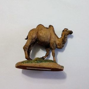 May include: A brown camel figurine with a hump on its back. The figurine is standing on a green and brown base.