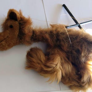 May include: A brown plush camel puppet with long fur and a long neck. The puppet is attached to a string and is lying on a white surface.