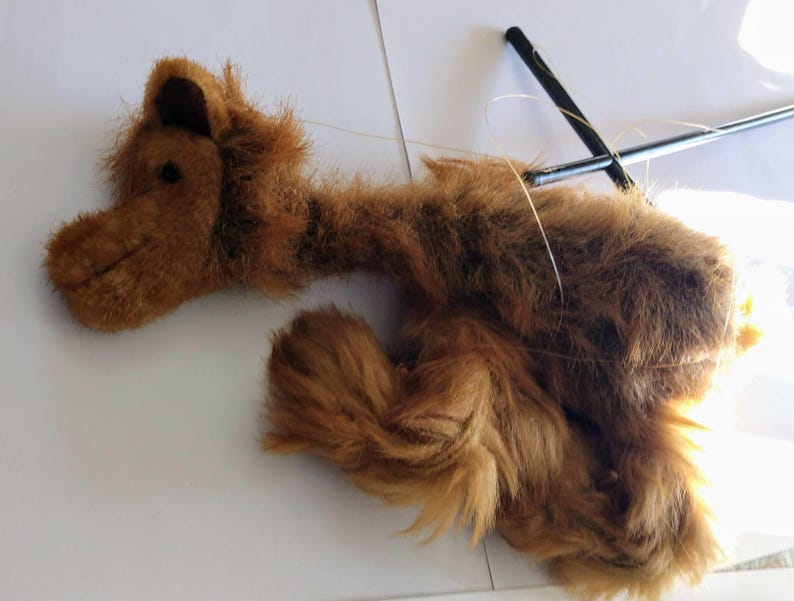 May include: A brown plush camel puppet with long fur and a long neck. The puppet is attached to a string and is lying on a white surface.