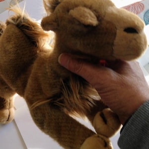 May include: A brown plush camel toy with a long neck and a fluffy mane. The camel is lying on its side with its head turned to the side. The camel has a soft, cuddly texture.