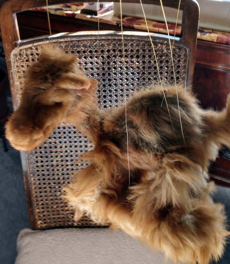 May include: A brown and black fur puppet hanging from strings on a wooden chair. The puppet has a long, furry tail and a long, furry neck.
