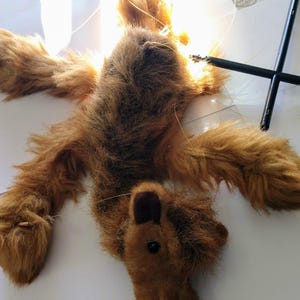 May include: A brown furry puppet of a dog with black eyes and a brown nose. The puppet is hanging from strings and is lying on a white surface.