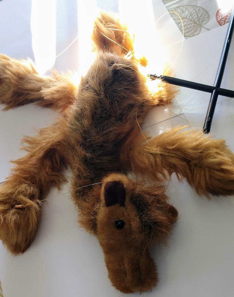 May include: A brown furry puppet of a dog with black eyes and a brown nose. The puppet is hanging from strings and is lying on a white surface.