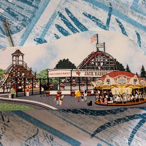 May include: A vintage postcard depicting a colorful amusement park scene with a wooden roller coaster called "Jack Rabbit" and a carousel. The postcard also features a smaller wooden roller coaster called "Fast-Fast" and a red train.