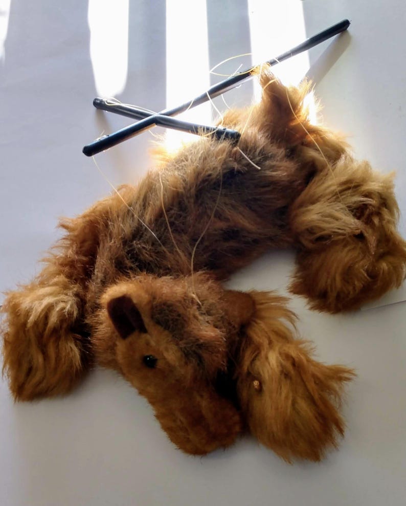 May include: A brown furry puppet of a dog with a long tail and a black control rod. The puppet is lying on its back with its legs in the air.