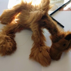 May include: A brown plush camel toy with long fur. The camel is lying on its side with its head turned to the side. The camel has a long neck and a small head with a black nose and eyes.