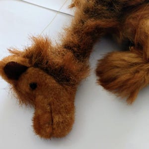 May include: A brown plush camel toy with a long neck and a long tail. The camel is lying on its side with its head turned to the side.