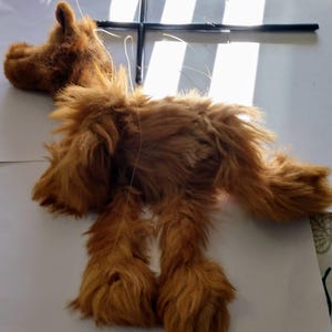 May include: A brown furry puppet of a dog with its legs and head hanging down. The puppet is attached to a black cross-shaped frame with strings.