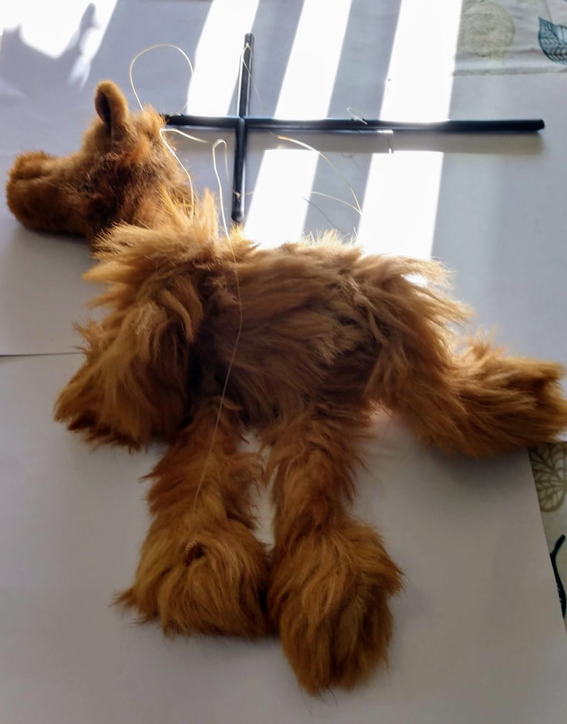 May include: A brown furry puppet of a dog with its legs and head hanging down. The puppet is attached to a black cross-shaped frame with strings.