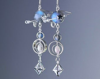 Puede incluir: Pendientes de plata con temática celestial. Cada pendiente presenta una cuenta redonda azul e iridiscente, un dije de estrella y un diseño geométrico con un colgante de cristal.