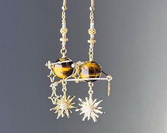 May include: Gold earrings with tiger's eye stone spheres and star-shaped charms. The earrings are suspended from a gold chain with small beads.
