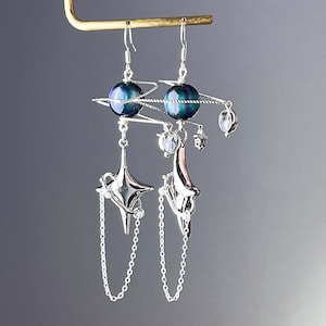 Può includere: Un paio di orecchini d'argento con un ciondolo a forma di stella e sfera. La sfera è blu e verde iridescente. Gli orecchini sono collegati da una catena d'argento.