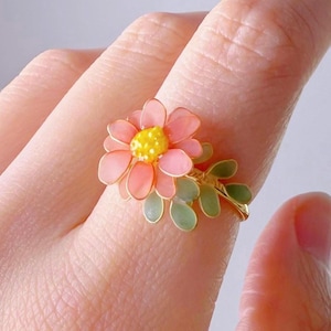 Puede incluir: Un delicado anillo con una flor rosa con un centro amarillo y hojas verdes. El anillo está hecho de metal y está diseñado para parecer una flor real.