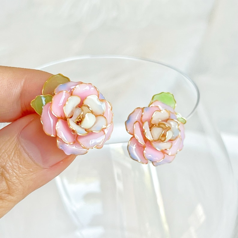 May include: A pair of floral stud earrings. Each earring features a pink, white, and light blue flower with gold-colored accents and green leaves. The earrings are set against a soft, blurred background.