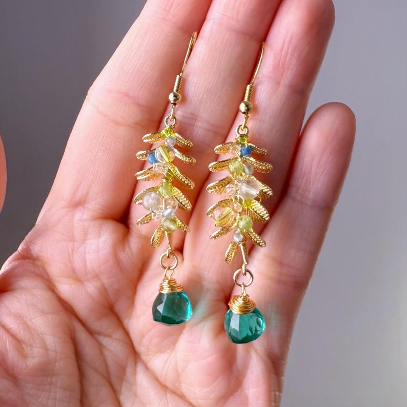 May include: A pair of gold-colored earrings with a leaf-like design. The earrings feature small, colorful beads in shades of yellow, green, and blue, and a teardrop-shaped, teal-colored gemstone at the bottom. The earrings are held in a hand.