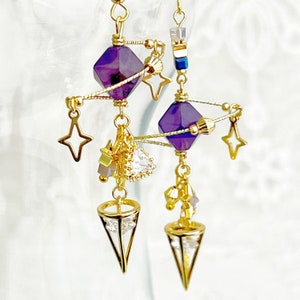 May include: Purple and gold dangle earrings with a star, crystal, and geometric design. The earrings feature a purple crystal, a gold star, and a gold geometric shape.