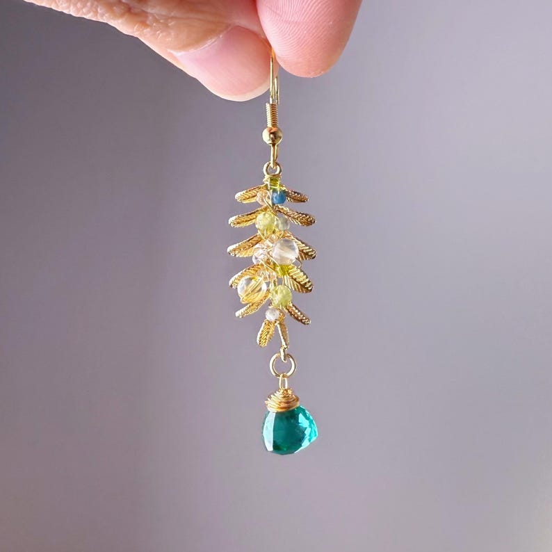 May include: A gold-colored earring featuring a cascading design with small, colorful beads and a teardrop-shaped, teal-colored gemstone at the bottom. The earring has a delicate, floral-inspired structure, with a hook for easy wear.