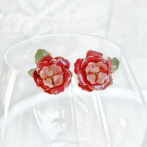 May include: A pair of floral stud earrings. Each earring features a red and pink flower design with gold-colored accents. The earrings have green leaf details. The earrings are displayed on a clear glass surface.