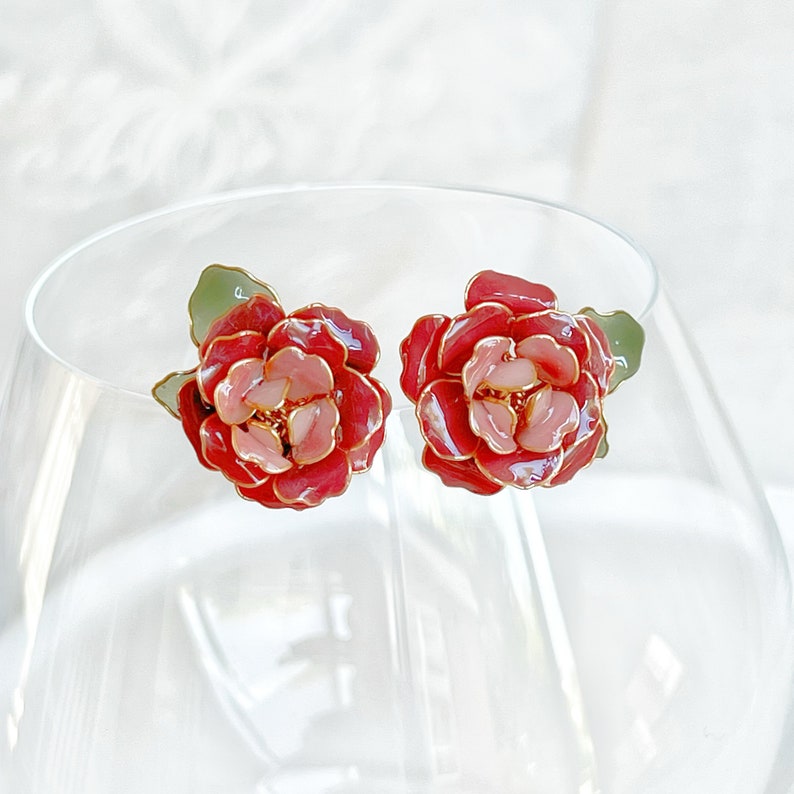 May include: A pair of floral stud earrings. Each earring features a red and pink flower design with gold-colored accents. The earrings have green leaf details. The earrings are displayed on a clear glass surface.