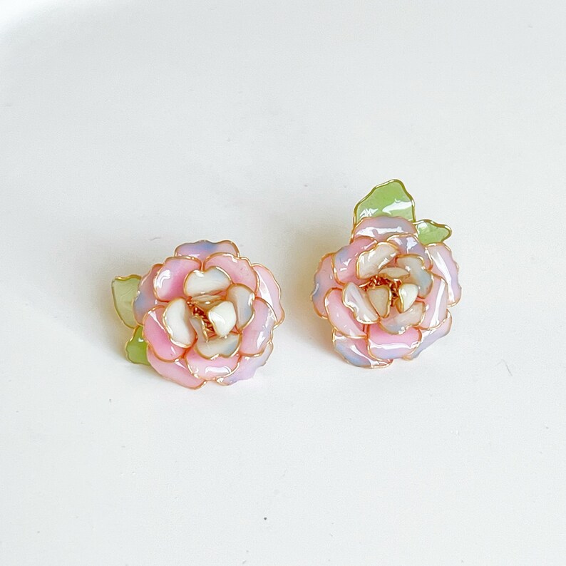 May include: A pair of floral stud earrings. Each earring features a pink, white, and light blue flower design with gold-colored accents. The earrings have a green leaf detail. The earrings are set against a white background.