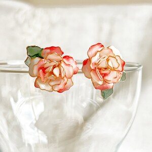 May include: A pair of floral stud earrings. Each earring features a delicate, layered flower design with white, pink, and red petals, outlined in gold. Green leaves accent the base of each flower. The earrings are set against a blurred background.