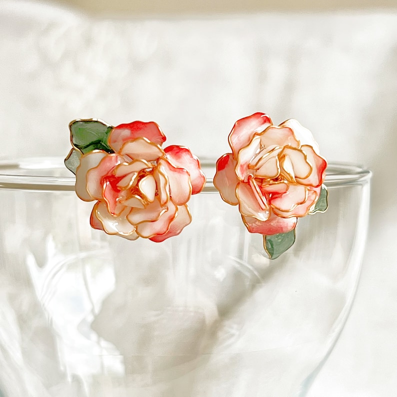 May include: A pair of floral stud earrings. Each earring features a delicate, layered flower design with white, pink, and red petals, outlined in gold. Green leaves accent the base of each flower. The earrings are set against a blurred background.