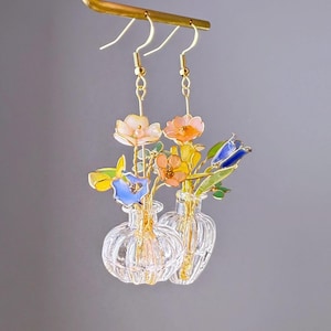 May include: A pair of gold earrings with glass vases and colorful resin flowers. The flowers are pink, yellow, blue, and green.