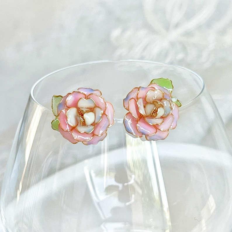 May include: A pair of floral stud earrings. Each earring features a pink, white, and light blue rose design with gold-colored trim and a green leaf. The earrings are set against a soft, blurred background.