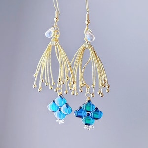 Puede incluir: Un par de pendientes colgantes con alambre dorado y cuentas de vidrio azules y transparentes. Los pendientes tienen un diseño floral con un grupo de pétalos de vidrio azules en la parte inferior. La parte superior de los pendientes tiene una cuenta transparente en forma de lágrima.