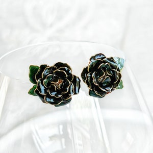 May include: A pair of dark green floral stud earrings with gold-colored trim. The earrings are shaped like roses and have a glossy finish. The earrings are set against a soft, white background.