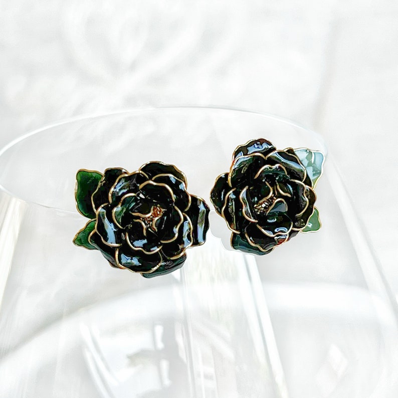 May include: A pair of dark green floral stud earrings with gold-colored trim. The earrings are shaped like roses and have a glossy finish. The earrings are set against a soft, white background.