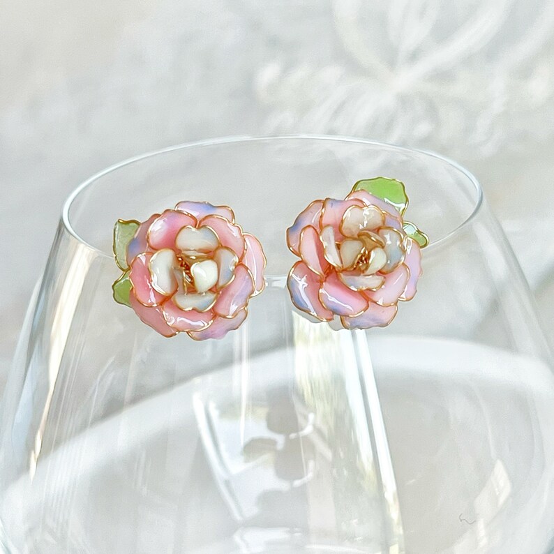May include: A pair of floral stud earrings. Each earring features a pink, white, and lavender flower design with gold-colored accents. The earrings have green leaves and are set against a blurred background.