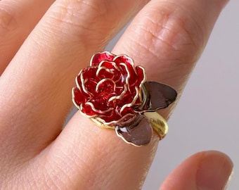 May include: A gold ring with a red rose design. The rose is made of metal and has a gold finish. The leaves are also made of metal and have a dark brown finish.