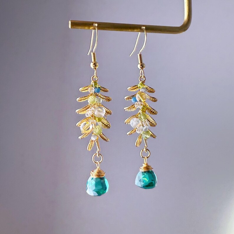 May include: A pair of gold-colored dangle earrings. Each earring features a gold-colored leaf design with small, clear, green, and blue beads. The earrings end with a faceted, teal-colored teardrop-shaped bead.