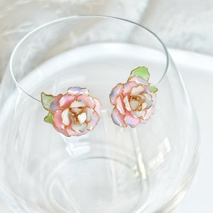 May include: A pair of floral stud earrings. Each earring features a pink, white, and light blue flower with gold-colored trim and a green leaf. The earrings are displayed in a clear glass.