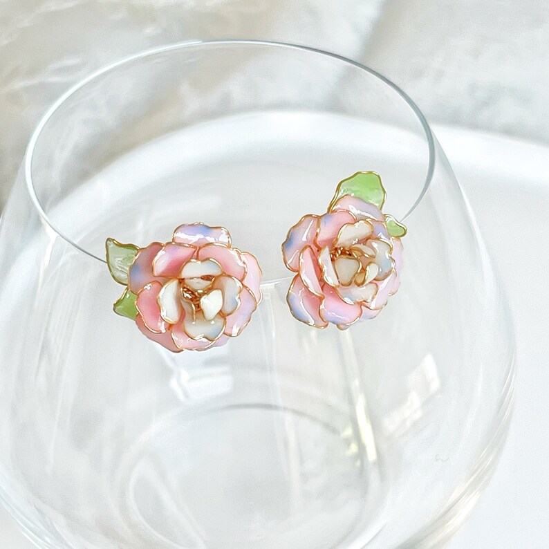 May include: A pair of floral stud earrings. Each earring features a pink, white, and light blue flower with gold-colored trim and a green leaf. The earrings are displayed in a clear glass.