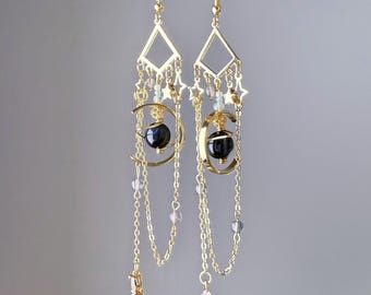 May include: Gold-toned dangle earrings featuring a diamond-shaped top, stars, and a crescent moon design. Each earring has a black sphere suspended within the moon, with delicate chains and dangling charms. The earrings are approximately 3 inches long.