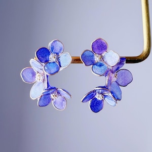Könnte beinhalten: Blumenohrringe mit blauen und violetten Blütenblättern und goldfarbenen Akzenten. Jede Blume hat eine kleine, weiße Perle in der Mitte. Die Ohrringe werden vor einem weichen, farbverlaufenden Hintergrund präsentiert.