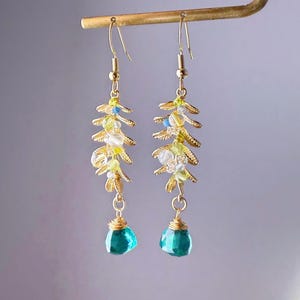 May include: Gold-toned dangle earrings featuring a leaf design with small yellow, white, and blue beads. Each earring has a faceted teal teardrop-shaped bead at the bottom. The earrings are suspended from a gold bar.