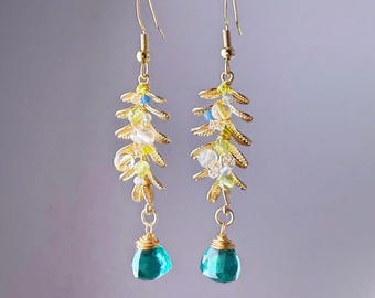 May include: Gold-toned dangle earrings featuring a leaf design with small yellow, white, and blue beads. Each earring has a faceted teal teardrop-shaped bead at the bottom. The earrings are suspended from a gold bar.