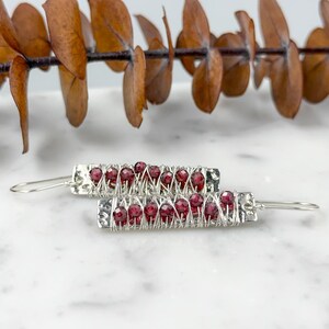 May include: A pair of silver earrings with red garnet beads wrapped in wire. The earrings are dangle style and have a hammered texture.