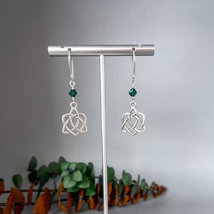 May include: Silver Celtic knot earrings with a green crystal dangle. The earrings are on a display stand.