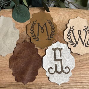 Puede incluir: Cinco posavasos de cuero en varios tonos de marrón y crema, dispuestos sobre una superficie de madera. Tres posavasos presentan una letra con un diseño de corona de laurel. Los otros dos posavasos son lisos.