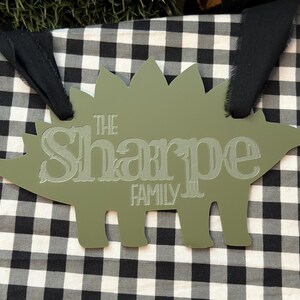 Op de afbeelding: Een olijfgroen dinosaurusvormig bord met de woorden "THE Sharpe FAMILY" gegraveerd in een lichtere tint. Het bord heeft een zwart lint om op te hangen en is geplaatst tegen een zwart-witte geruite achtergrond.