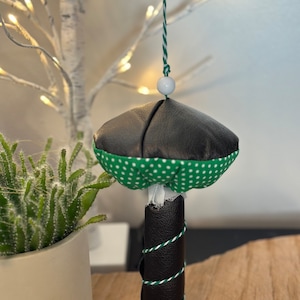 Puede incluir: Un adorno en forma de hongo negro y verde con lunares blancos que cuelga de una cuerda blanca y verde. El adorno está hecho de tela y tiene una base de cuero negro.