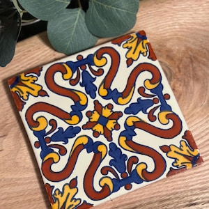 May include: Square ceramic tile with a cream background and a repeating pattern of red, yellow, and blue floral and scroll designs. The tile is placed on a light wood surface, with green leaves in the background.