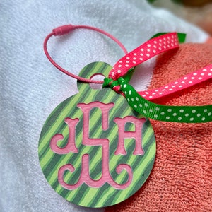 May include: A green and pink monogrammed luggage tag with the initials "JLA" in a script font. The tag is attached to a pink and white striped towel with a pink and white polka dot ribbon and a green and white polka dot ribbon.