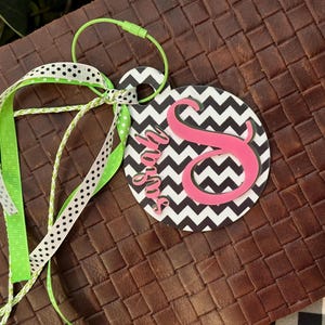 Könnte beinhalten: Ein schwarzes und weißes Chevron-Muster-Etikett mit dem Namen "Sarah" und einem pinkfarbenen Buchstaben "S" in kursiver Schrift. Das Etikett ist an einem grün-weißen Band und einer grünen Schnur befestigt.